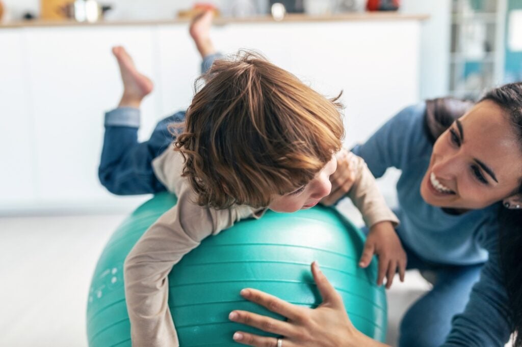 Shot of young beautiful mother playing with fitness ball on the floor with her son looking at camera in living room at home.